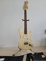 Full front view of cream-colored Fender Squier Stratocaster electric guitar on stand with bag laid on table, showing overall wear and condition