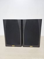 Front view of the JAMO 38 speakers with grills attached, showing black fabric covers and small gold brand badges at the bottom.