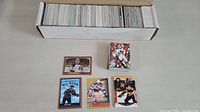 Box of over 600 hockey and football cards along with 5 individual cards displayed in front showing a range of players and conditions.