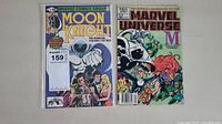 Photo showing two comic books side by side. Left is Moon Knight #1, right is The Official Handbook of the Marvel Universe #7 with Moon Knight on the cover.