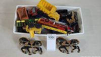 Photo showing two black metal horse figurines and multiple toy trucks including a yellow dump truck, a red fire truck with ladder, and other colored trucks all in a white tray.
