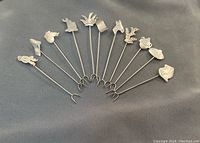 11 Mexican sterling silver cocktail picks arranged in a fan shape on dark fabric, showing all unique designs on each pick's top.