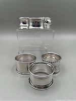Six sterling silver napkin rings shown with two in front and four stacked on clear display block behind. Polished cylindrical form, some with slight edge detailing.