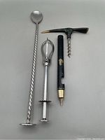 Three bar tools shown laid out on a white surface: a twisted silver plated muddler with flat base, a silver plated pickle/olive tongs, and a brass opener/corkscrew/ice pick designed as an ice axe.