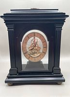 Full view of the Bombay Company mantle clock showing black wooden case with side columns, stepped top, brass feet and glass front revealing skeletonized clock face.