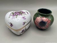Heart shaped white fine china trinket box painted with purple Victorian Violets flowers and gold accents next to a small rounded green Moorcroft vase with pink and purple Anemone flower design.