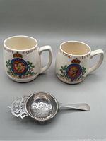 Three items pictured: two ceramic mugs and one silver plate tea strainer arranged on a neutral background.