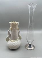 Two vases shown side by side: white Belleek dolphin spill vase with scalloped top and two dolphin handles, and tall clear etched glass vase with sterling base.