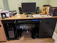 Front view of wood veneer desk showing three drawers on the right, pull-out keyboard tray, computer tower space on left, monitor on top with speakers and lamp
