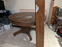 Photo of the round oak table with the 10 inch leaf stored vertically, showing the table top surface with wear and need for refinishing.