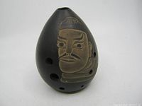 Front view of vintage black clay xun flute with carved human face and finger holes visible.