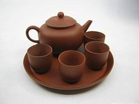 Front angle view of the Yixing clay mini teapot set with four cups arranged on the round tray, showing intact teapot and cups, two cups exhibit damage.