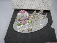 Glass figurine on an oval glass base showing two horses decorated with red accents pulling a rounded carriage with pink and yellow dots and green leaves, alongside glass flowers. Original box with Crystal World branding visible in the background.