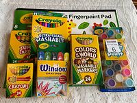 Photo of all new Crayola products in colorful packaging on a white surface including crayons, jumbo crayons, washable markers, window crayons, washable paints, and fingerpaint pad.