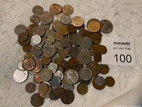 Top view of a large assortment of coins spread on a beige cloth surface. Various sizes and tones from silver to copper with different designs and wear conditions.