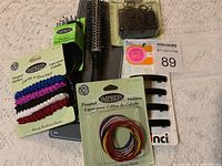 Photo showing the batch of new hair accessories including elastic hair bands, two types of hair brushes, brown hair elastics, and colored ponytail holders arranged on a light fabric background.