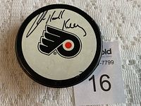Black hockey puck with white top displaying the Philadelphia Flyers logo and a signature believed to be Bob Kelly's