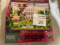 Photo showing unopened 1000 piece Hometown Collection puzzle box with colorful scene featuring hot air balloons, buildings, and bunny-themed displays.