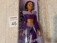 Vintage Disney Princess Jasmine doll shown inside a clear cylindrical plastic container with purple and pink Disney Princess branding. Doll has midriff exposed purple outfit and black hair. Container top lid missing.