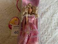 Full view of Sleeping Beauty doll in original clear plastic packaging with pink background, showing doll wearing pink dress with long sleeves and ruffled collar, blonde curly hair visible.