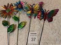 Four metal yard decorations including two colorful flowers on stems with leaves and two colorful butterflies on stems.