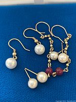 Close-up of four pairs of earrings on blue fabric, showing gold hooks and pearls with red beads