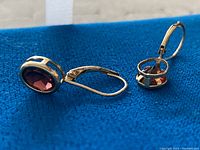 Close-up side view of two 10K gold drop earrings showing oval red stones and lever-back clasps on blue fabric background.