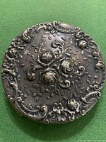 Top view of antique silver compact mirror showing detailed embossed floral and scroll design.
