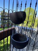 Photo showing two black hanging flower baskets with chains attached, hanging on an outdoor iron railing with visible green grass and buildings in the background.