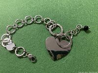 Silver chain bracelet with circular links, heart charm, and a black bead detail on a green surface.