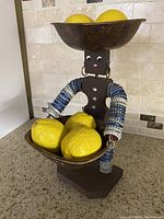 Folk art figure with lemons in upper and lower wooden bowls, showing bottle cap decorated arms and metal face.