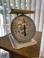 Front angle photo of vintage metal kitchen scale with decorated dial face and flat weighing platform on top.