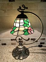 Lamp fully lit showing colorful stained glass shade with pink flower and green leaves pattern and curved metal arm with detailed metal base.