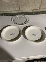 View of the three pie plates: two white ceramic CorningWare Cornflower pattern plates and one clear glass plate on a white surface.