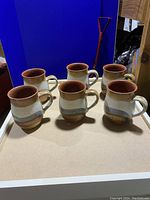 Six handmade pottery mugs arranged on a board with visible exterior paint design and handles on the right side.