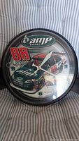 Front view of 10.5 inch round battery operated wall clock showing Dale Earnhardt NASCAR racing car on the face with AMP Energy logo and number 88, along with signature text. Clock hands are visible and the clock is enclosed in a black frame.