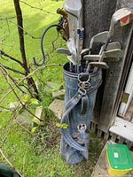 Full view of golf bag with assorted golf clubs outside on a concrete step near wooden wall and greenery.