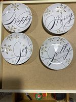 Top view of four different holiday dessert plates with words 'Happy', 'Merry', 'Joy', and 'Jolly' in cursive and decorated with gold snowflake patterns.