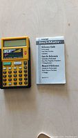Front view of yellow and black InchMate+ calculator and instruction manual on a wooden surface.