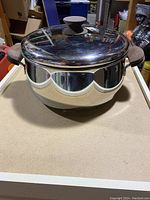 Stainless steel cooking pot with lid shown from the top and front angle, displaying the reflective surface and dark knob on lid.