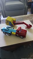 Photo showing red and teal vintage metal truck and yellow and red digger side by side on a wooden table, displaying their condition and color details.