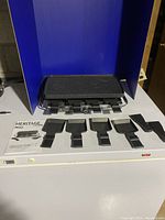 Black reversible party grill with 8 raclette trays and 8 scrapers displayed in front, all placed on a white surface with a blue backdrop. Shows condition and accessories.