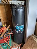 Photo showing a 100lb black punching bag with 'Round One' logo, stored upright in a corner with surrounding tools and equipment visible.