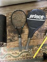 Two adult tennis rackets side by side on wooden surface with Prince Pro Oversize cover on one.