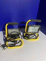 Two yellow framed portable utility work lights with black power cords, shown powered off on a light surface against a blue background.