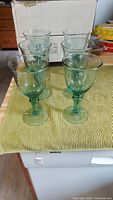 Front view of 6 green-blue glass wine goblets arranged in two rows on a green cloth on washing machine, showing their size and shape.