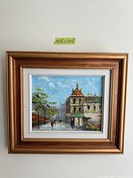 Framed oil painting showing a vintage city street scene with people walking, trees, and buildings. The painting has a gold ornate frame and white mat with dimensions approximately 15.5 x 13.5 inches.