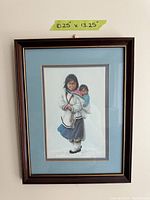Framed art print showing a woman in traditional Inuit clothing carrying a child on her back, with dimensions 10.25 x 13.25 inches noted on tape.