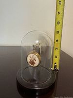 Pressed flower charm pendant displayed under a glass dome with black base, shown next to a tape measure indicating about 4 inches height.
