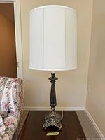 Photo of a vintage table lamp with white cylindrical fabric shade and ornate dark metal base with silver detailing on claw feet, placed on a wooden surface next to floral fabric couch.